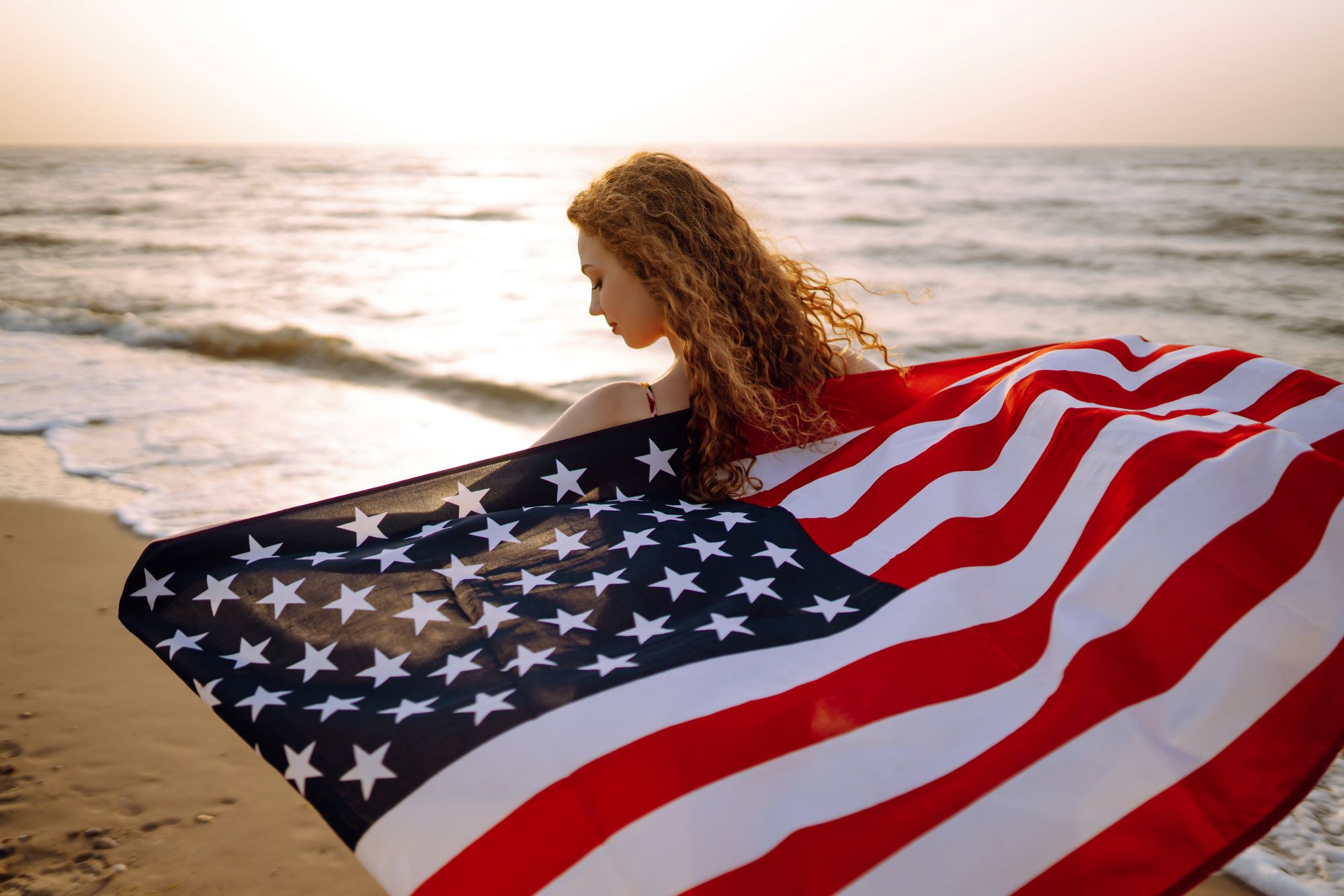 Young woman with american flag on the beach at sunset.